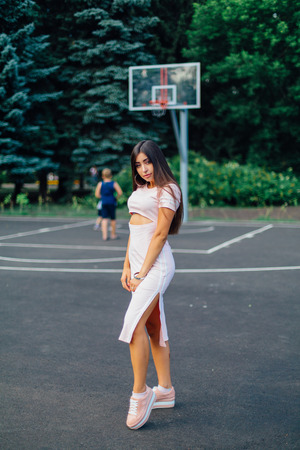 Charming brunette female dressed in a pink dress posing on the basketball court during sunset.の写真素材