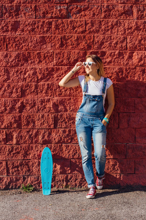 Portrait of a smiling woman dressed in overalls and sunglasses standing with her skateboard next to the red wall.の写真素材