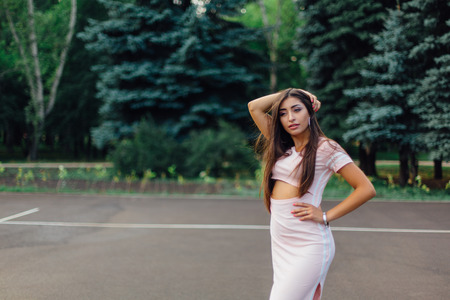 Charming brunette female dressed in a pink dress posing on the basketball court during sunset.の写真素材