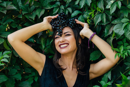Portrait of young sexy brunette in black dress with black grapes on head.の写真素材