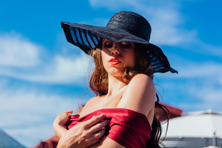 Beautiful young woman in red dress and balack hat in summer sunny day on the blue sky background. Retro fashionの写真素材