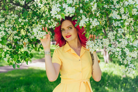 Beautiful and young woman with bright red hair and ref lips standing next to a blooming apple tree in a yellow dress. Spring portrait.の写真素材