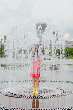 Young wet pretty girl with two braids in yellow boots and with transparent umbrella stands inside of fountain. Rainy day in city.の写真素材