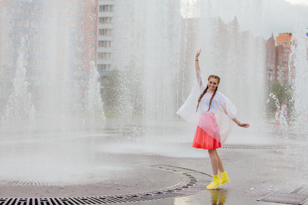 Young wet pretty girl with two braids in yellow boots stands near fountain. Rainy day in city.の写真素材