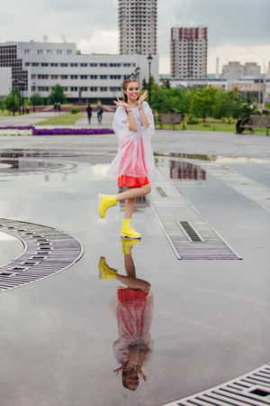 Young wet pretty girl with two braids in yellow boots stands near fountain. Rainy day in city. Young girl reflecting in the wet groundの写真素材