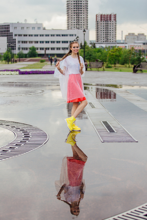 Young wet pretty girl with two braids in yellow boots stands near fountain. Rainy day in city. Young girl reflecting in the wet groundの写真素材