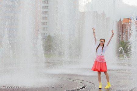 Young wet pretty girl with two braids in yellow boots stands near fountain. Rainy day in city.の写真素材