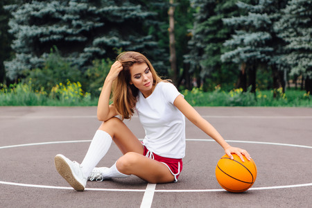 Beautiful young blonde girl dressed in white t-shirt, shorts and sneakers, sits on a basketball court with ball outdoors. Copy space.の写真素材
