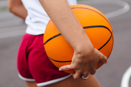 Young woman dressed in white t-shirt, shorts holding a ball on a basketball court outdoors. Close up woman's fanny in shortsの写真素材