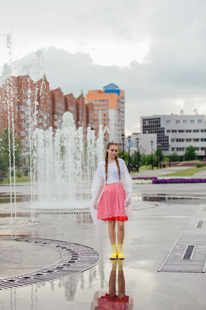 Young wet pretty girl with two braids in yellow boots stands near fountain. Rainy day in city.の写真素材