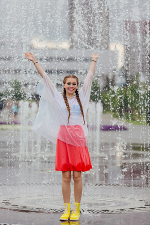 Young wet pretty girl with two braids in yellow boots and with transparent rain clothe stands inside of fountain. Rainy day in city.の写真素材