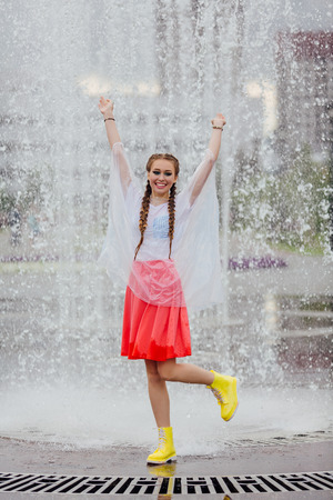 Young wet pretty girl with two braids in yellow boots and with transparent rain clothe stands inside of fountain. Rainy day in city.の写真素材