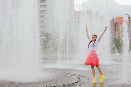 Young wet pretty girl with two braids in yellow boots stands near fountain. Rainy day in city.の写真素材