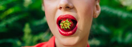 Beautiful young pin up girl dressed in red shirt with bright make up and fresh natural strawberry in mouth. Close up.の写真素材