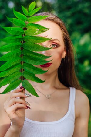Portrait of female with beautiful green eyes dressed in whire shirt with a leaf of fern near one eye in a summer park.の写真素材
