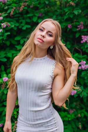 Spring portrait of a charming blond woman wearing beautiful white dress standing next to blooming purple lilac bush.の写真素材