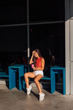 Stylish happy young woman wearing white shorts and sneakers holding pinc cup of coffee to go....