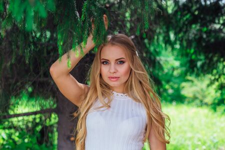 Spring portrait of a charming blond woman wearing beautiful white dress standing next under fur tree.の写真素材