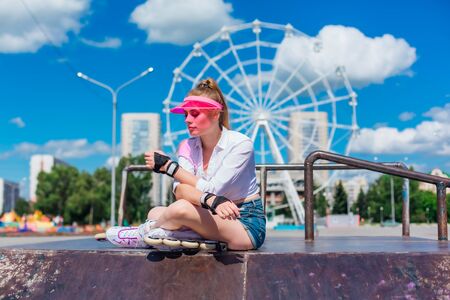 Portrait of an emotional girl in a pink cap visor wearing protective gloves and roller skate sitting on the background of ferris wheel and relaxing.の写真素材