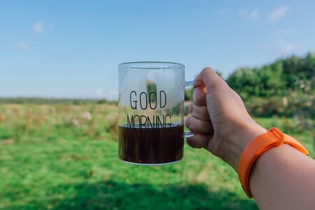 Right hand of woman holding cup of black hot Americano coffee. Clear glass with words GOOD MORNING on a green field background. Drink conceptの写真素材