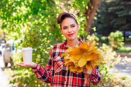 Beautiful woman with make up and hair in pin up style holding big bouquet of maple yellow leaves and...