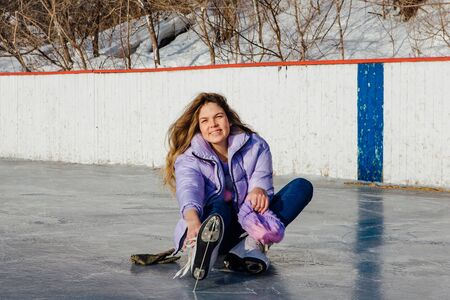 Lovely young woman sitting on ice ring and tieing shoelaces. Girl is going to skating on ice in a...