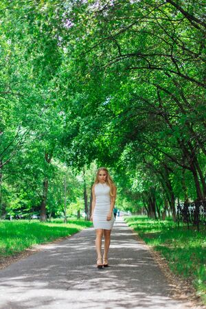 Spring portrait of a charming blond woman wearing beautiful white dress walking on the road under green trees.の写真素材