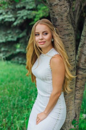 Spring portrait of a charming blond woman wearing beautiful white dress standing on grass next to rowan tree in a park.の写真素材