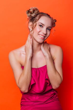 The girl in a red dress on a orange background in the studio. Blonde girl with two hair knots holding hands on shoulders, looking to the camera and smiling.の写真素材
