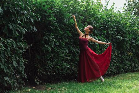 Woman ballerina in red ballet dress dancing in pointe shoes in autumn park next to the wall of bushes. Ballerina standing in beautiful ballet poseの写真素材