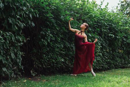 Woman ballerina in red ballet dress dancing in pointe shoes in autumn park next to the wall of bushes. Ballerina standing in beautiful ballet poseの写真素材