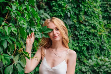 Portrait of a young beautiful woman with make up and curly blond hair, holding grape leaf close to the face. Copy space.の写真素材