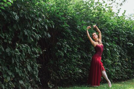 Woman ballerina in red ballet dress dancing in pointe shoes in autumn park next to the wall of bushes. Ballerina standing in beautiful ballet poseの写真素材
