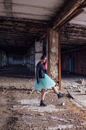 Portrait of a young girl with pink hair dressed in leather jacket and a blue skirt standing inside of collapsed building surrounded by ruinsの写真素材