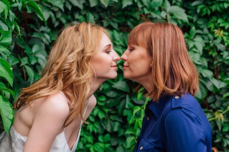 Beautiful mother and her grown up daughter standing nose to nose to each other on a summer day outside on a green nature backgroundの写真素材