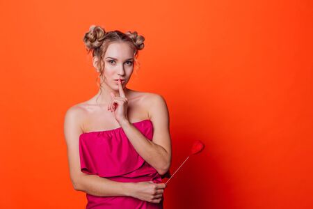 The girl in a red dress on a orange background in the studio. Blonde girl with two hair knots showing shhhhh sign, holding red heart on the stick.の写真素材