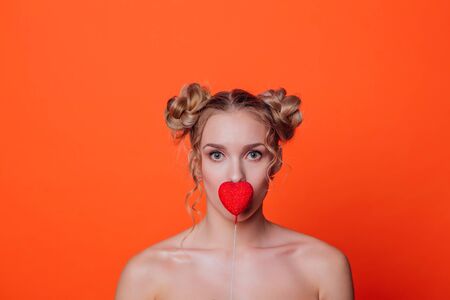 The girl on a orange background in the studio. Blonde girl with two hair knots holding red heart on the stick close to the lips.の写真素材
