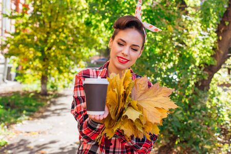 Beautiful woman with make up and hair in pin up style holding big bouquet of maple yellow leaves and black cup of coffee to go in a sunny autumn day. Copy space.の写真素材