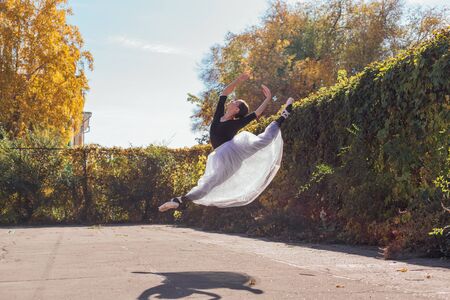 Woman ballerina in a white ballet skirt dancing in pointe shoes in a golden autumn park. Ballerina jumping doing a splitの写真素材