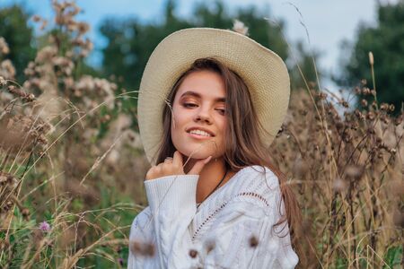 Beautiful brunette teenage girl dressed in a white sweater, jeans and cowboy straw hat sitting in dry brown bur grass, smiling and laughing during sunsetの写真素材