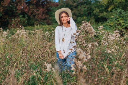Beautiful brunette teenage girl dressed in a white sweater, jeans and cowboy straw hat standing in dry brown bur grass, smiling and laughing during sunsetの写真素材