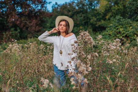 Beautiful brunette teenage girl dressed in a white sweater, jeans and cowboy straw hat standing in dry brown bur grass, smiling and laughing during sunsetの写真素材