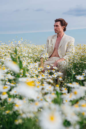 Tall handsome man dressed in a white suit on on naked body sitting on a white chair in camomile flowers fieldの写真素材