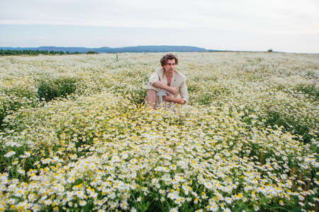 Tall handsome man dressed in a white suit on naked body sitting on a white chair in camomile flowers fieldの写真素材