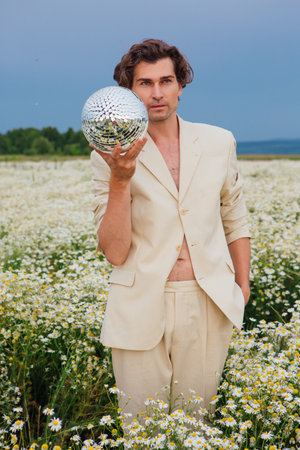 Tall handsome man dressed in a white suit on naked body standing in camomile flowers field, holding mirror disco ball.の写真素材