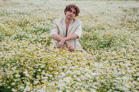 Tall handsome man dressed in a white suit on naked body sitting on a white chair in camomile flowers fieldの写真素材
