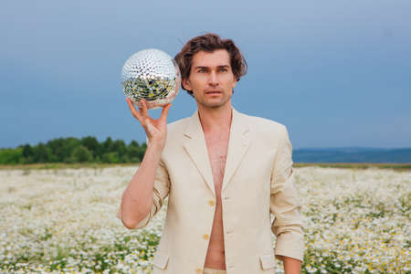 Tall handsome man dressed in a white suit on naked body standing in camomile flowers field, holding mirror disco ball.の写真素材