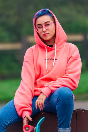 Young girl with short multicolored hair and nose piercing dressed in pink hoodie sits with skateboard in skatepark during morning fog. Portrait of a tomboy with plastic skateboardの写真素材