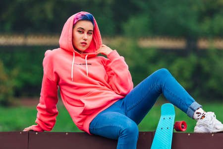 Young girl with short multicolored hair and nose piercing dressed in pink hoodie sits with skateboard in skatepark during morning fog. Portrait of a tomboy with plastic skateboardの写真素材