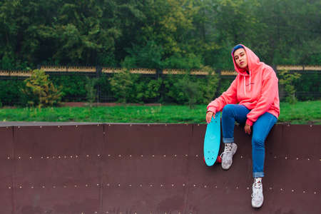 Young girl with short multicolored hair and nose piercing dressed in pink hoodie sitting with skateboard in skatepark during morning fog. Portrait of a tomboy with plastic skateboardの写真素材